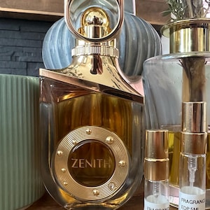 Könnte beinhalten: Ein goldfarbener Parfümflakon mit dem Wort "ZENITH" auf der Vorderseite. Der Flakon hat ein einzigartiges Design mit einem kreisförmigen Akzent und einem goldenen Ring oben. Zwei kleine Duftprobenflaschen sind ebenfalls sichtbar.