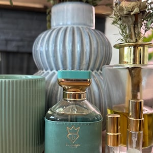 Op de afbeelding: Een glazen parfumfles met een teal lederen omhulsel, een goudkleurige dop en een teal deksel, gelabeld Bvlgari. Daarnaast staan twee kleine doorzichtige proefflesjes met gouden doppen, gelabeld Fragrance EDP 3ML en Fragrance EDP 5ML.