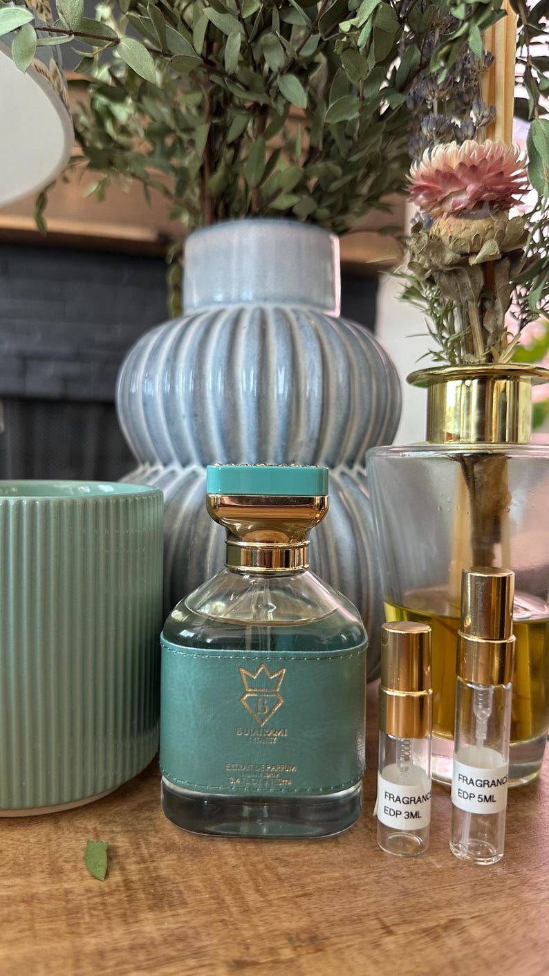 Op de afbeelding: Een glazen parfumfles met een teal lederen omhulsel, een goudkleurige dop en een teal deksel, gelabeld Bvlgari. Daarnaast staan twee kleine doorzichtige proefflesjes met gouden doppen, gelabeld Fragrance EDP 3ML en Fragrance EDP 5ML.