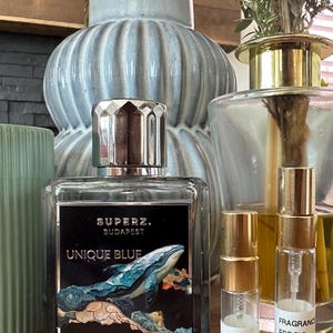Op de afbeelding: Een rechthoekige glazen parfumfles met een zilveren dop en een zwart etiket met een walvisillustratie en de tekst SUPERZ. BUDAPEST UNIQUE BLUE, naast twee kleine doorzichtige glazen verstuivers met het label FRAGRANCE EDP 3ML en 5ML.
