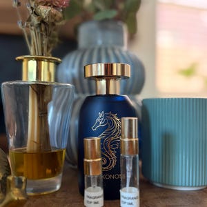 May include: A dark blue matte perfume bottle with a gold seahorse graphic and the text KONOS, accompanied by two small clear glass sample vials with gold caps, each featuring a white label with black text, positioned in front of a vase.