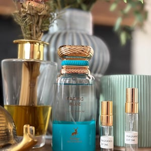 May include: A collection of perfume bottles and samples. The main bottle is clear with a gold cap and a turquoise base, labeled "PACIFIC BLUE". Two smaller sample bottles with gold caps are also present. The background includes vases and dried flowers.