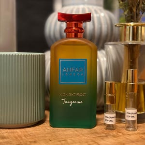 May include: A rectangular perfume bottle with a gradient finish transitioning from orange at the top to green at the bottom, featuring a red cap and a blue label with the text ANFAP LONDON MIDNIGHT FROST Tangerine, alongside two small sample vials.