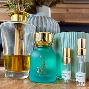 Könnte beinhalten: Ein türkisfarbener, mattierter Glasparfümflakon mit goldenem Verschluss und goldenem Leopardenmotiv, beschriftet mit MYKONOS, sowie zwei kleine, durchsichtige Glasprobenfläschchen mit goldenen Sprühköpfen und weißen Etiketten für 3ml und 5ml.