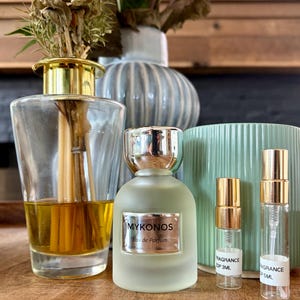Könnte beinhalten: Ein mattierter Glasparfümflakon mit der Aufschrift MYKONOS Eau de Parfum und einem goldenen Verschluss, begleitet von zwei kleinen, durchsichtigen Glasprobenfläschchen mit goldenen Sprühköpfen, beschriftet mit FRAGRANCE EDP 3ML und FRAGRANCE EDP 5ML.