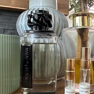 Op de afbeelding: Een rechthoekige glazen parfumfles met een zwarte dop en een zwart zijpaneel met het opschrift "LYNKED by AFNAN", naast twee kleine glazen proefflesjes met gouden verstuivers en witte labels met "FRAGRANCE EDP 3ML" en "FRAGRANCE EDP 5ML".