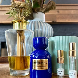Könnte beinhalten: Eine kobaltblaue Glasparfümflasche mit einem goldenen Etikett mit der Aufschrift INCEPTION Extrait de Parfum, neben zwei kleinen, durchsichtigen Glassprühfläschchen mit 3ml und 5ml Beschriftung, einem Glas-Raumduftdiffusor und einem hellgrünen Becher.