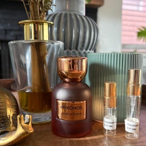 Könnte beinhalten: Eine matte braune Parfümflasche mit einem metallisch kupferfarbenen Verschluss und einem Etikett mit der Aufschrift MYKONOS Extrait de Parfum, begleitet von zwei kleinen, klaren Glassprühfläschchen mit der Aufschrift FRAGRANCE EDP 3ML und 5ML.