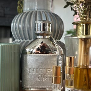 Może przedstawiać: Srebrny flakon perfum Nitro Pour Homme Elixir o teksturowanym metalicznym wykończeniu, w towarzystwie dwóch małych przezroczystych szklanych fiolek z próbkami ze złotymi nakrętkami, z etykietami Fragrance EDP 3ML oraz Fragrance EDP 5ML.