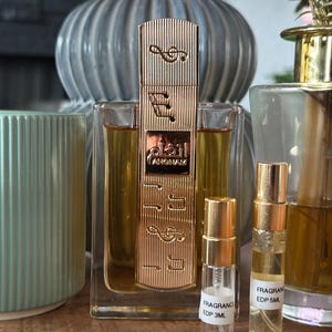 K&ouml;nnte beinhalten: Eine goldene Parf&uuml;mflasche mit Noten und arabischem Text, flankiert von zwei kleineren Probenflaschen. Die Parf&uuml;mflasche befindet sich in einem transparenten Glasbeh&auml;lter. Im Hintergrund befinden sich eine blaue Vase und ein gr&uuml;ner Kerzenhalter.