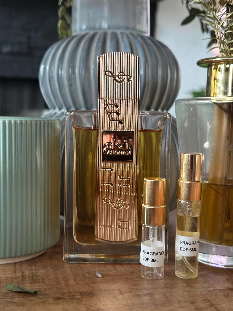 K&ouml;nnte beinhalten: Eine goldene Parf&uuml;mflasche mit Noten und arabischem Text, flankiert von zwei kleineren Probenflaschen. Die Parf&uuml;mflasche befindet sich in einem transparenten Glasbeh&auml;lter. Im Hintergrund befinden sich eine blaue Vase und ein gr&uuml;ner Kerzenhalter.