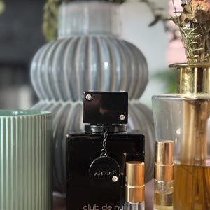 Puede incluir: Un frasco de perfume negro rectangular con el texto "club de nuit intense MAN" y dos muestras de fragancia más pequeñas. El frasco de perfume tiene una tapa negra con dos pequeños cristales. Las muestras tienen tapas doradas y etiquetas blancas.