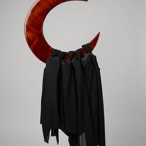 May include: A crescent moon-shaped wall hanging crafted from polished reddish-brown wood. Attached to the lower curve of the wooden moon are several strips of black fabric that hang vertically, creating a layered, draped effect across the structure.