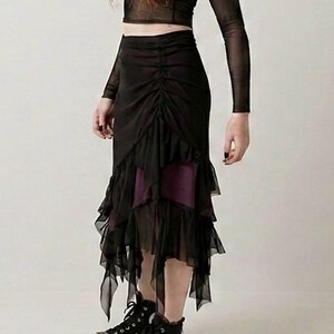 May include: A black sheer long-sleeve top paired with a black midi skirt featuring a ruched front panel, layered ruffled hem, and a dark purple underskirt. The outfit is completed with black canvas high-top trainers with white rubber soles.