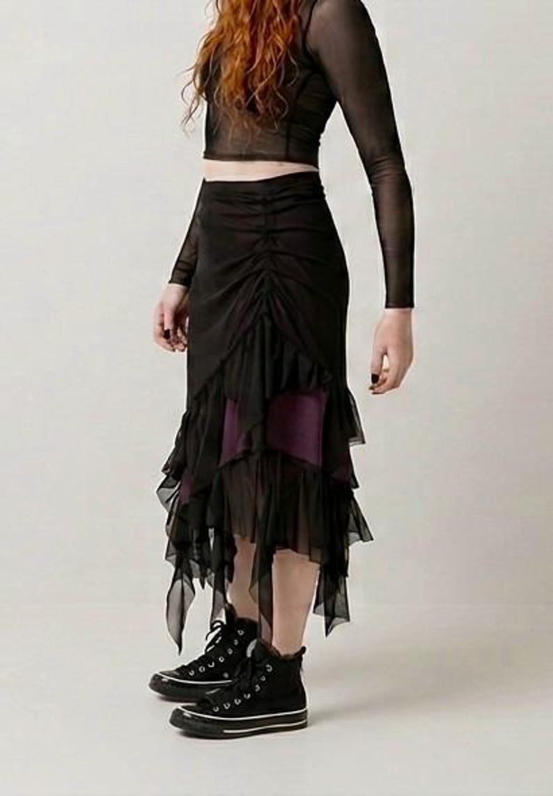 May include: A black sheer long-sleeve top paired with a black midi skirt featuring a ruched front panel, layered ruffled hem, and a dark purple underskirt. The outfit is completed with black canvas high-top trainers with white rubber soles.