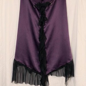 May include: A deep purple satin skirt featuring a vertical black ruffled trim down the centre, accented with two silver-toned metal rings at the top. The hemline is finished with a sheer black mesh ruffle that creates an asymmetrical layered effect.