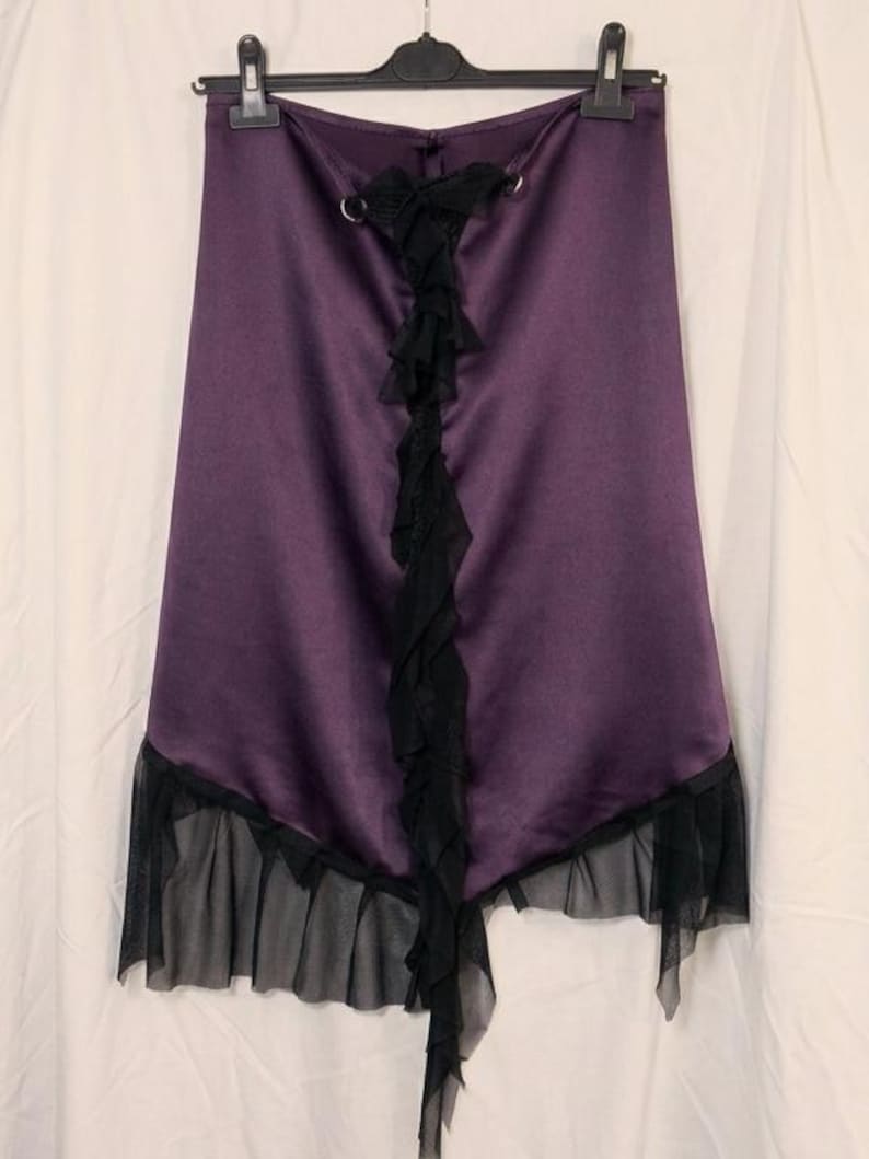 May include: A deep purple satin skirt featuring a vertical black ruffled trim down the centre, accented with two silver-toned metal rings at the top. The hemline is finished with a sheer black mesh ruffle that creates an asymmetrical layered effect.