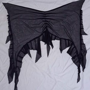 May include: A black sheer mesh wrap featuring a gathered centre design with vertical ruched detailing. The edges are finished with a ruffled trim and pointed, asymmetrical fabric panels that hang down from the sides and bottom hemline.