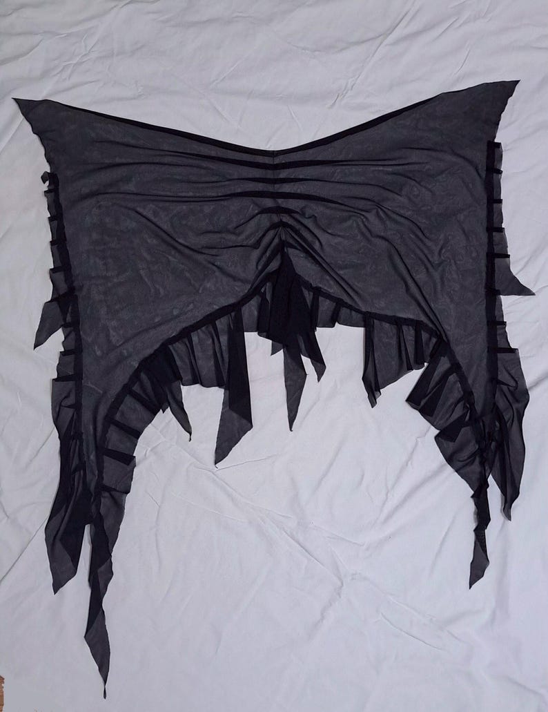 May include: A black sheer mesh wrap featuring a gathered centre design with vertical ruched detailing. The edges are finished with a ruffled trim and pointed, asymmetrical fabric panels that hang down from the sides and bottom hemline.