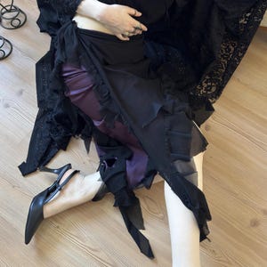 May include: A layered skirt featuring a deep purple satin base layer and a sheer black chiffon overlay with ruffled edges. The skirt is paired with a black lace top and black pointed-toe slingback heels with a thin stiletto heel.