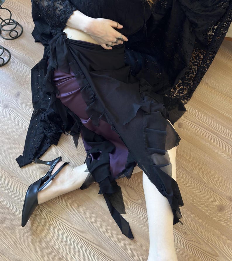 May include: A layered skirt featuring a deep purple satin base layer and a sheer black chiffon overlay with ruffled edges. The skirt is paired with a black lace top and black pointed-toe slingback heels with a thin stiletto heel.