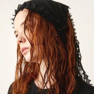 May include: A black headscarf featuring a ruffled edge along the front and a delicate lace trim that extends down the back. The fabric is lightweight and opaque, designed to be worn over the hair with a secure fit around the crown.