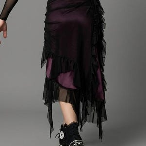 Puede incluir: Una falda de capas con una base de color morado oscuro y una capa superior de malla negra transparente con bordes con volantes. La falda tiene un dobladillo asimétrico con paneles de malla colgantes y se combina con zapatillas negras.