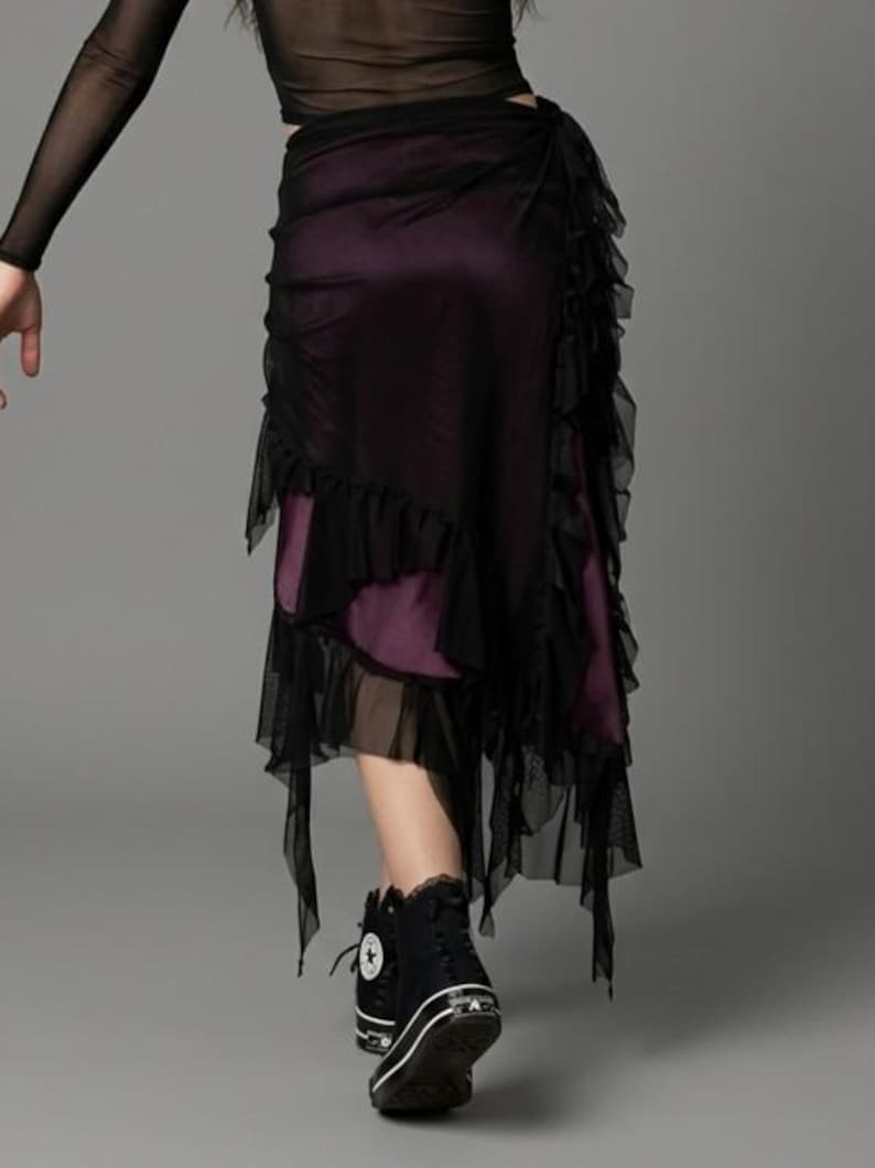 May include: A layered skirt featuring a deep purple base layer and a sheer black mesh overlay with ruffled edges. The skirt has an asymmetrical hemline with hanging mesh panels and is paired with black high-top trainers with white soles.