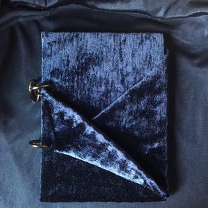 May include: A rectangular notebook covered in deep navy blue crushed velvet fabric. The cover features a folded corner detail and is bound on the left side with two circular gold-toned metal rings that secure the pages together.