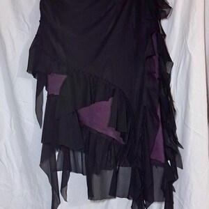 May include: A black skirt featuring layered sheer fabric panels and ruffled edges. The garment has an asymmetrical hemline and reveals a solid dark purple fabric layer underneath. The skirt is displayed on a black plastic hanger against a white background.