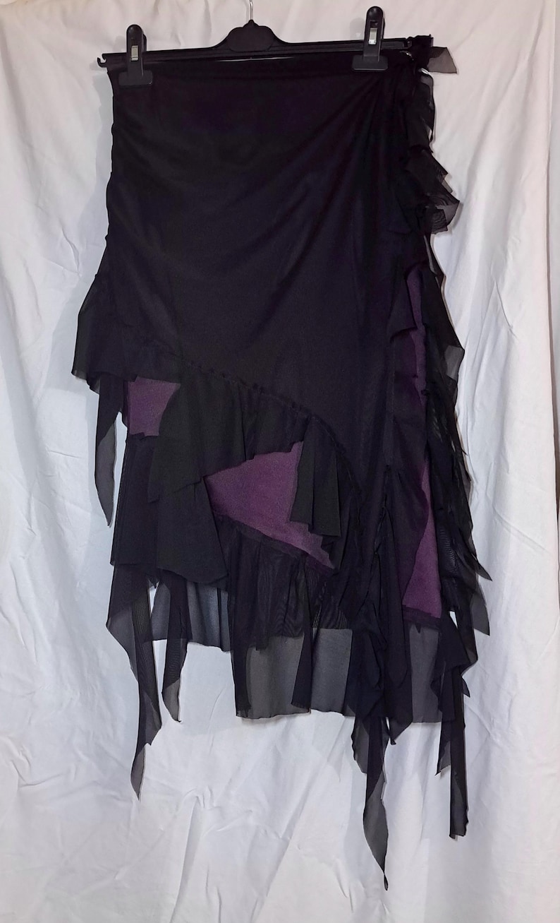 May include: A black skirt featuring layered sheer fabric panels and ruffled edges. The garment has an asymmetrical hemline and reveals a solid dark purple fabric layer underneath. The skirt is displayed on a black plastic hanger against a white background.