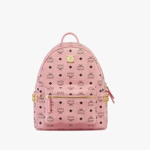 May include: Pink backpack featuring a repeating pattern of "MCM" letters and diamond shapes. The backpack has gold-coloured zips and hardware, a top handle, and two side pockets. The bag is approximately 25 cm tall.
