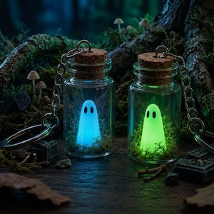 May include: Two clear glass bottle keychains with cork stoppers, each containing a small glowing ghost figure nestled in green moss. One ghost emits a bright blue light, while the other emits a vibrant green light, both featuring two black oval eyes.
