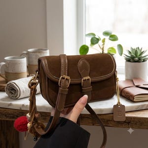 May include: A dark brown suede crossbody bag featuring a fold-over flap with two vertical buckled straps, gold-toned hardware, and a decorative leather charm with the letter E attached to the side, along with a dangling tassel of beads.