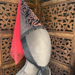 May include: A tall, conical headdress featuring a black fabric base with a repeating orange coral-like pattern. The base of the cone is trimmed with black braided cord, and a sheer red veil hangs from the back. A black chin strap secures the piece.