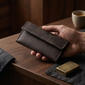 May include: A dark brown leather wallet with a flap closure. The wallet is held in a hand and sits on a wooden surface. A brass lighter rests nearby. The wallet appears to be made of high-quality leather and has visible stitching.