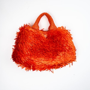 May include: A bright orange handbag featuring a dense, shaggy fringe texture covering the entire exterior. The bag includes two woven top handles and a small triangular metal logo plaque centered near the top opening of the bag.