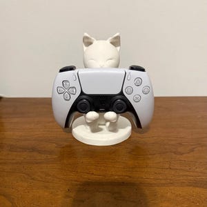 May include: A white cat-shaped controller stand holding a white and black game controller. The stand is made of ceramic or a similar material. The controller has buttons and directional pads. The stand is on a wooden surface.