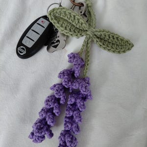 May include: A crochet keychain featuring two purple lavender sprigs with green leaves attached to a metal ring. A black Nissan car key fob and a silver-toned metal tag with the text "every dog needs a home" and a heart shape are also attached.