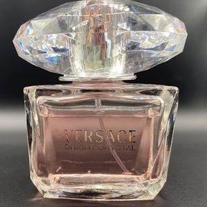 May include: A square glass perfume bottle containing pale pink liquid, featuring the text VERSACE BRIGHT CRYSTAL on the front. The bottle is topped with a large, clear, faceted crystal-shaped stopper that resembles a diamond in its design.