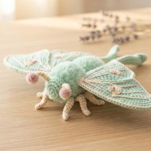 May include: A crocheted moth figurine featuring a fuzzy mint green body, two small pink pom-pom antennae, and wide, flat wings in mint green with cream-colored embroidered details. The moth has six cream-colored crocheted legs extending outward.
