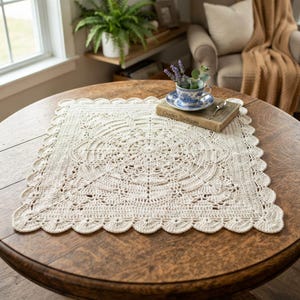 May include: A square, off-white crochet doily featuring an intricate, circular lace pattern in the center that transitions into a scalloped edge. The doily is placed on a round wooden table, with a book titled Crochet Traditions resting on it.