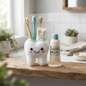 May include: A white crocheted tooth-shaped holder with black eyes and pink cheeks, containing two toothbrushes and a tube of toothpaste. Beside it stands a crocheted beige toothpaste tube with the text TOOTH PASTE embroidered in black.
