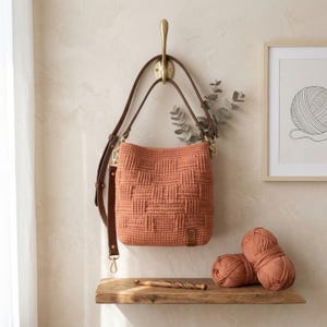 May include: A terracotta-colored crochet handbag featuring a textured basketweave pattern, a brown leather adjustable shoulder strap with gold-toned metal hardware, and a small rectangular leather brand tag attached to the lower right corner.
