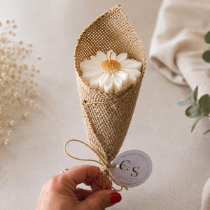 Könnte beinhalten: Eine weiße gänseblümchenförmige Kerze, eingewickelt in einen beigen Jute-Kegel. Die Kerze hat eine gelbe Mitte und einen einzelnen Docht. Ein kleines rundes Etikett mit dem Text "GRACIAS POR VENIR" und den Buchstaben "CS" ist mit einer Schnur befestigt.