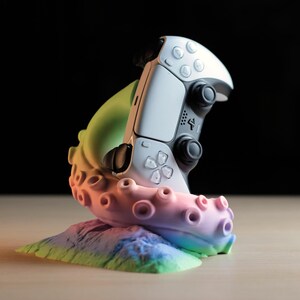 Könnte beinhalten: Ein weiß-schwarzer Videospiel-Controller, der von einem mehrfarbigen, im Farbverlauf gestalteten Oktopus-Tentakel-Ständer gehalten wird. Der Tentakel hat eine glatte Textur mit sichtbaren Saugnäpfen und verläuft von Grün zu Rosa und Blau.