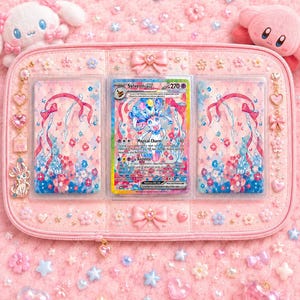 Puede incluir: Un tarjetero rosa con una carta Pokémon Sylveon EX en el centro. El soporte está decorado con lazos, corazones y adornos florales. Se ven peluches de un conejito blanco y un Kirby rosa.