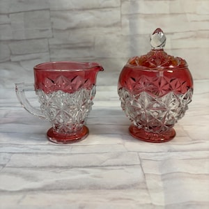 Könnte beinhalten: Ein Milchkännchen und eine Zuckerdose mit Deckel aus Glas in einer Farbkombination aus Rot und Transparent. Beide Gefäße weisen im unteren Bereich ein facettiertes Rautenmuster auf, während der obere Rand mit einem glatten roten Band verziert ist.