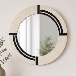 Op de afbeelding: Een ronde spiegel met een licht houten frame. Het frame heeft zwarte accenten in een geometrisch patroon. Het reflecterende oppervlak van de spiegel wordt omgeven door licht hout, wat een modern, decoratief stuk creëert.