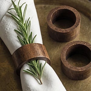 May include: Three cylindrical napkin rings crafted from dark brown wood with a visible grain pattern. One ring secures a white fabric napkin folded with a fresh sprig of green rosemary tucked inside, while two additional rings sit nearby.